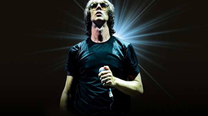 Image of Richard Ashcroft in a black t-shirt wearing sunglasses and holding a microphone, set against a static black background backlit by a bright white spotlight